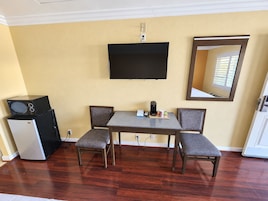 Una habitación con televisión de pantalla plana en la pared, una kitchenette pequeña con refrigerador y estufa, una mesa de comedor con dos sillas y un espejo en la pared.