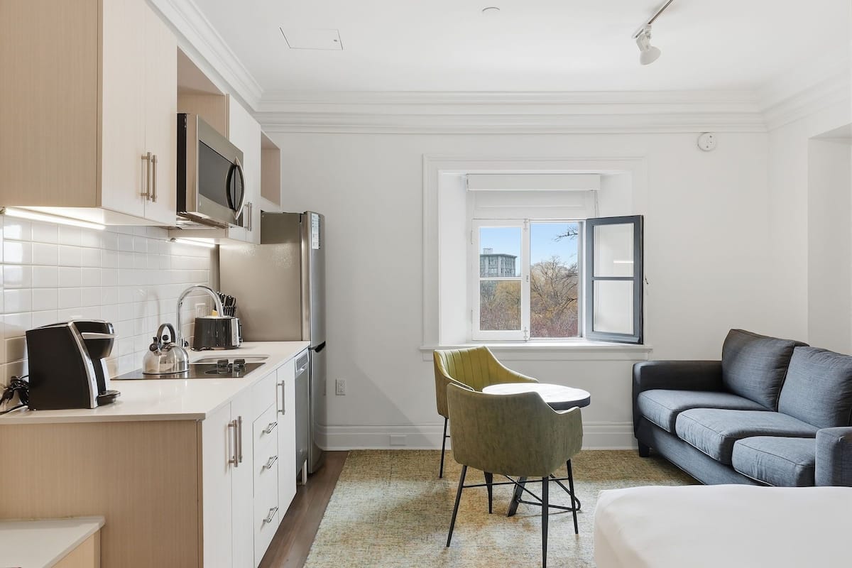 A modern kitchen with white cabinets, a microwave, and a coffee machine. There is a dining area with a table and chairs, and a gray sofa.