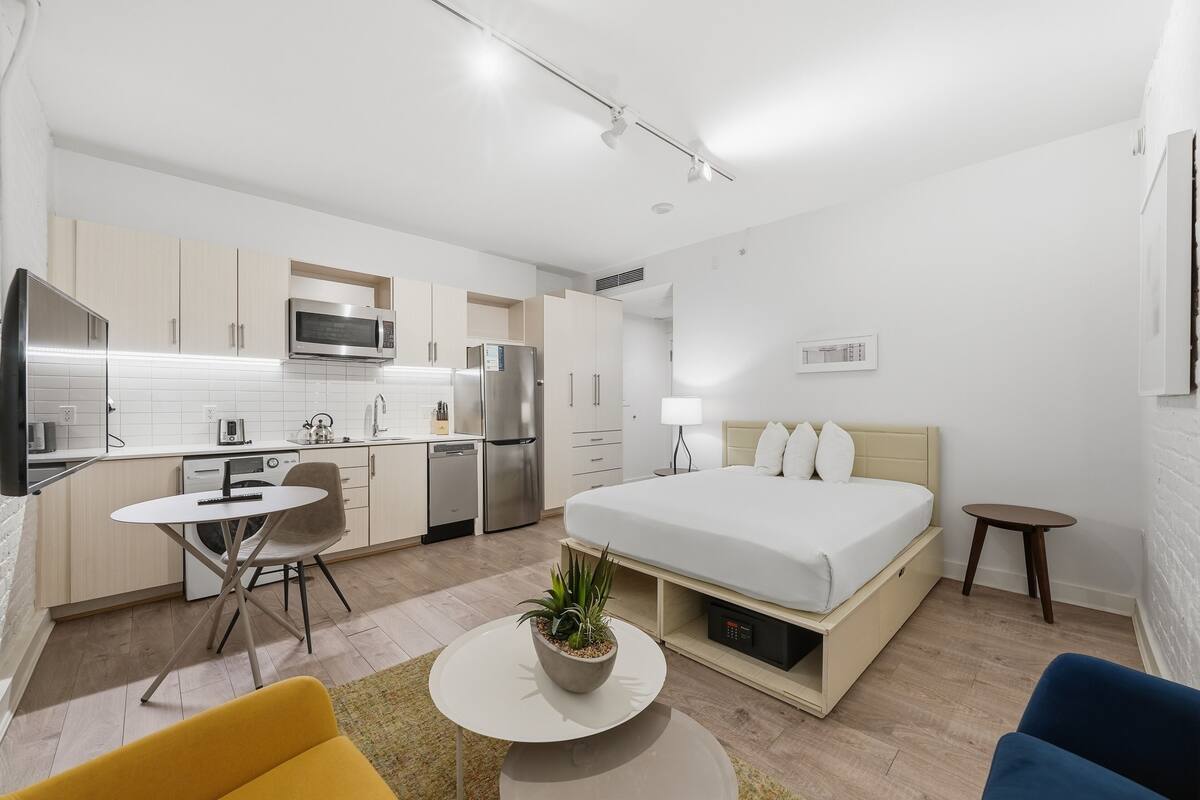 A modern hotel room with a bed, a small dining table, a kitchen area, and a living area with a sofa.