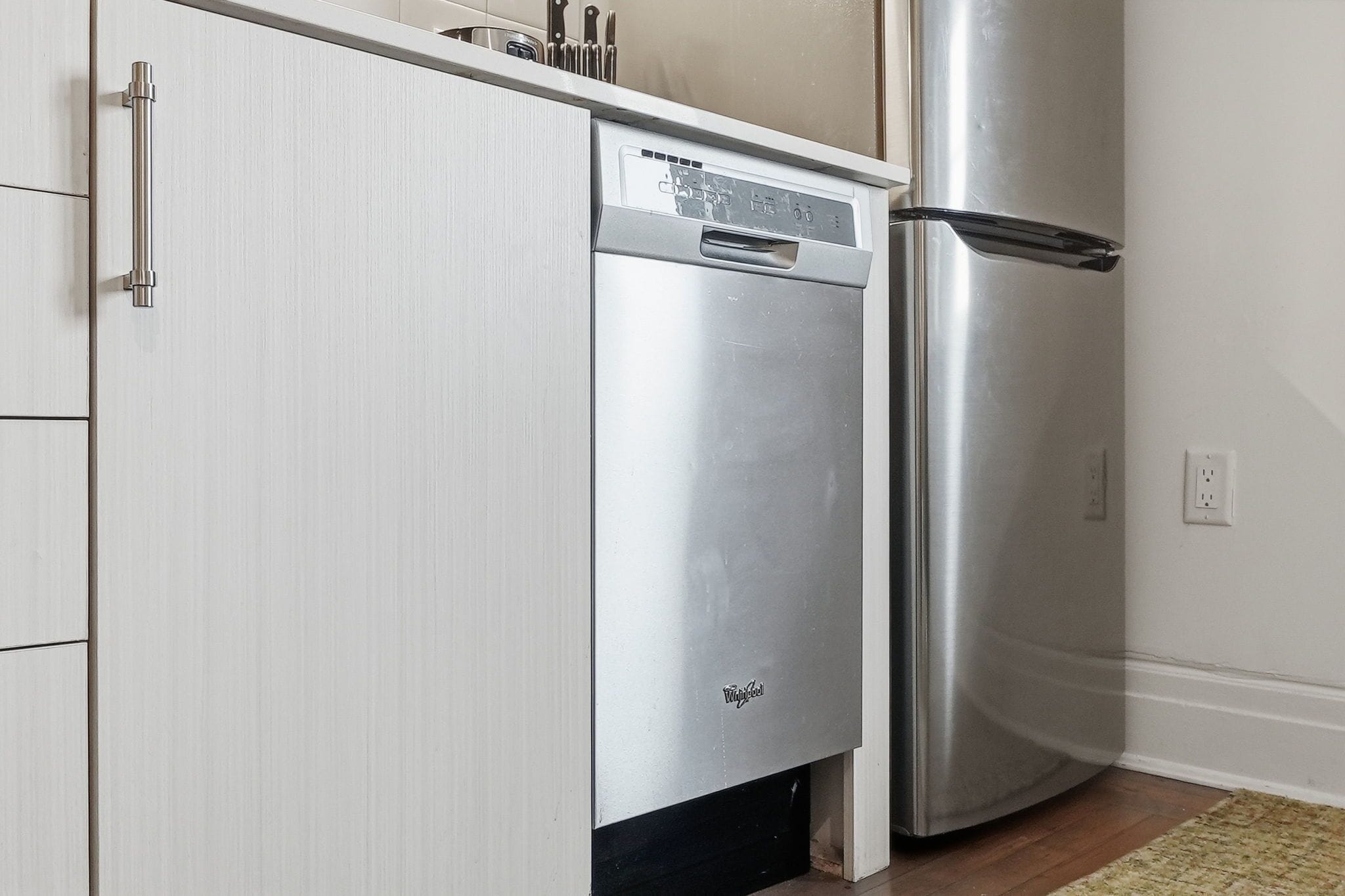 A stainless steel refrigerator and dishwasher in a kitchen with white cabinetry and a wooden floor.