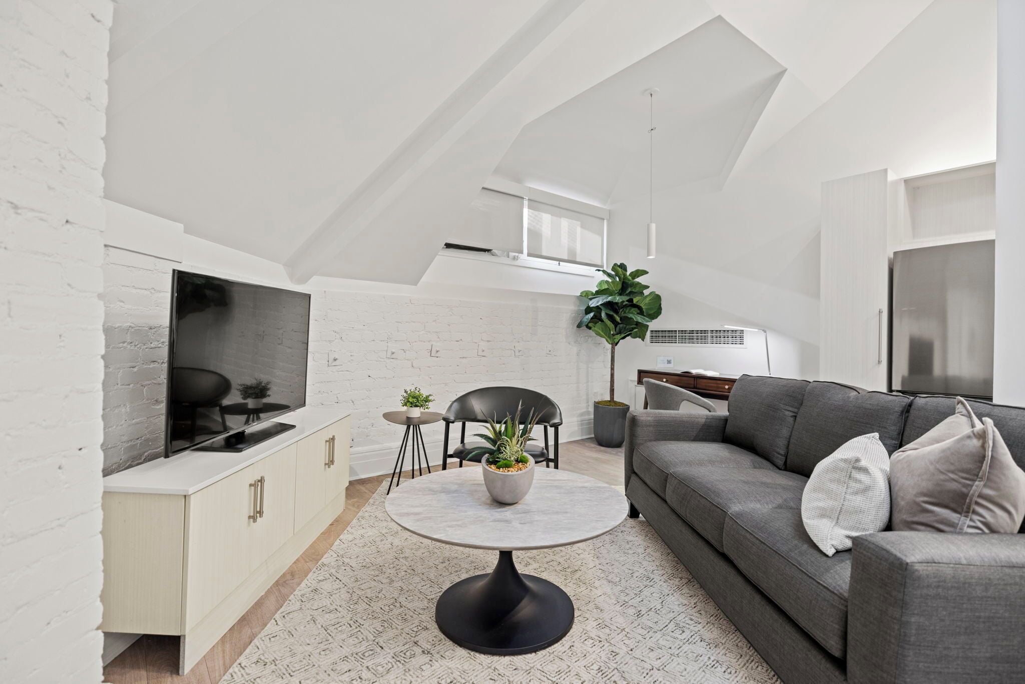 Ein modernes Wohnzimmer mit einer weißen Ziegelwand, einem grauen Sofa, einem runden Couchtisch und einem Fernseher auf einem weißen Schrank.