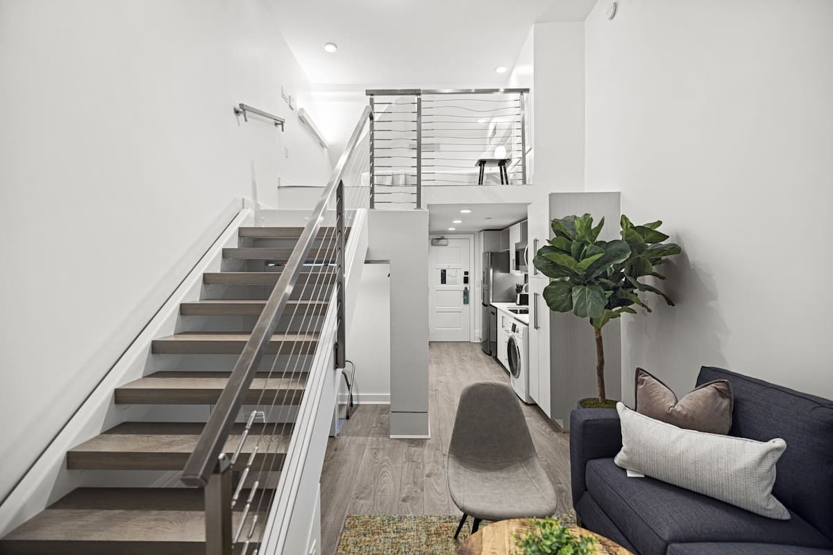 A modern interior with a staircase leading to a second floor, a kitchen area, and a seating area with a sofa and cushions.