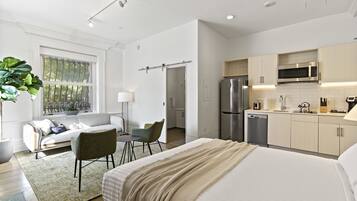 A modern hotel room with a large bed, a seating area with a sofa and chairs, a kitchenette with a refrigerator and microwave, and a large window with a view of trees.