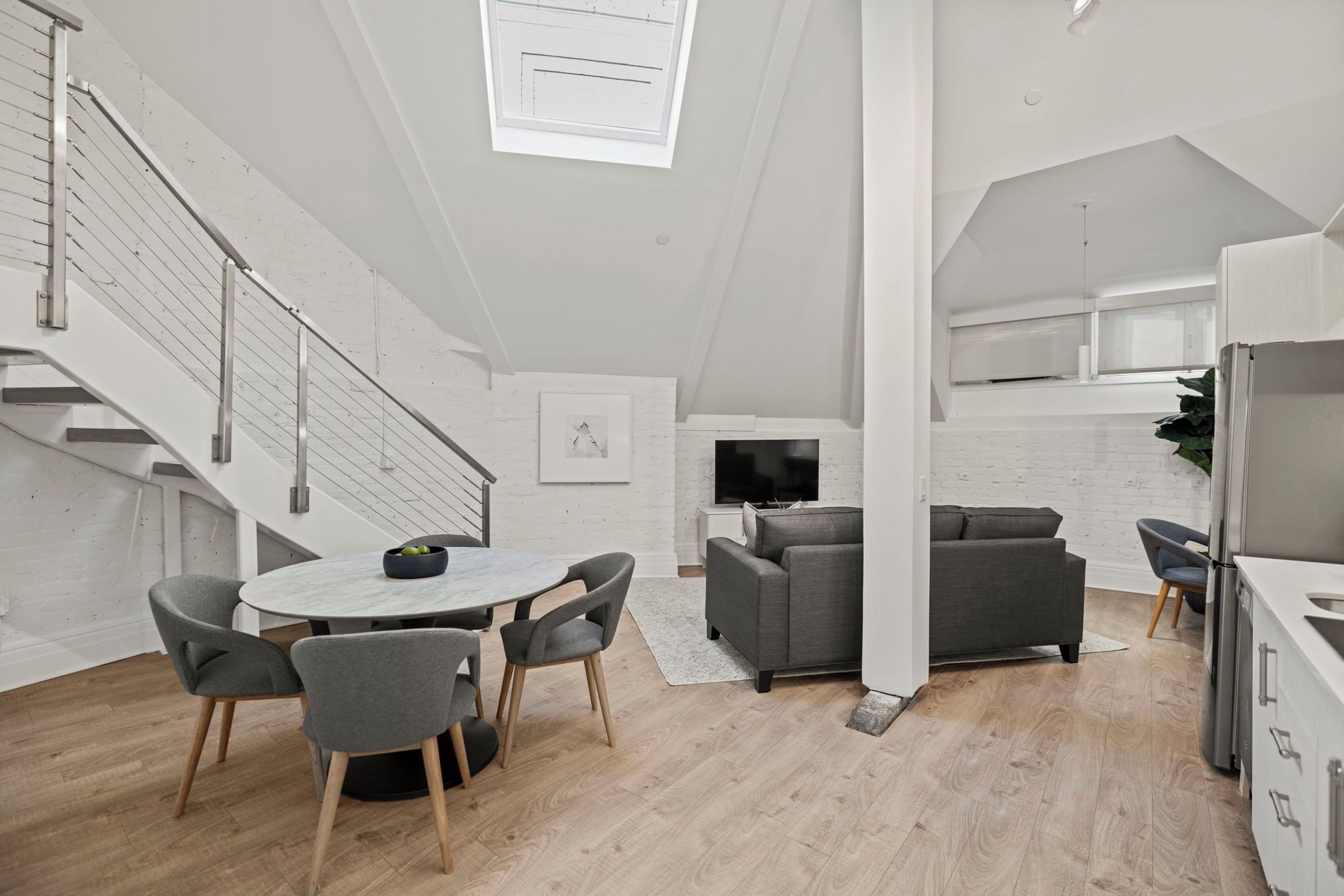 A modern kitchen with a built-in oven, a dining area with a round table and chairs, a grey sofa, and a staircase with metal railings.
