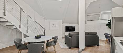 A modern kitchen with a built-in oven, a dining area with a round table and chairs, a grey sofa, and a staircase with metal railings.