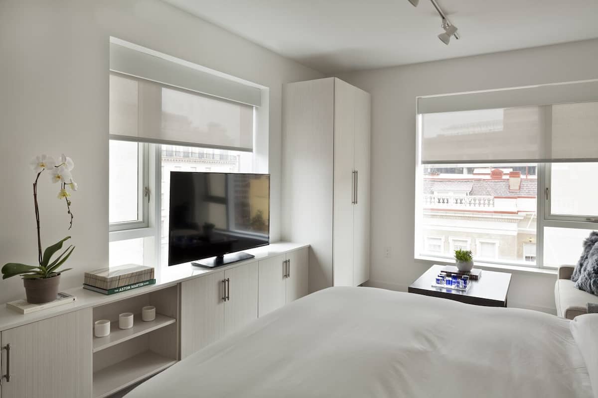 A modern bedroom with a bed, a TV on a stand, a window with blinds, and a view of buildings outside.