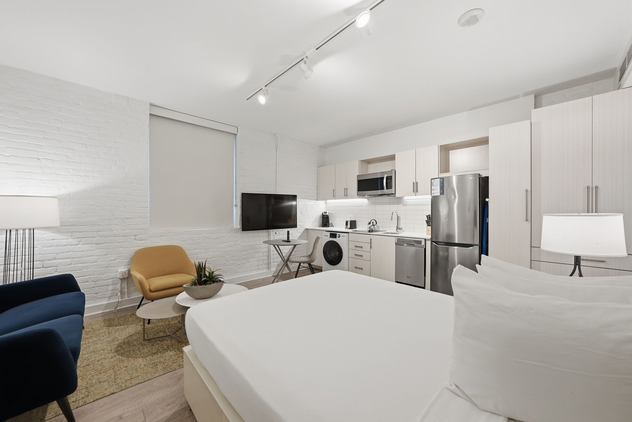 A modern hotel room with a bed, a sofa, a small table with a plant, a TV, a microwave, a refrigerator, and a washing machine.