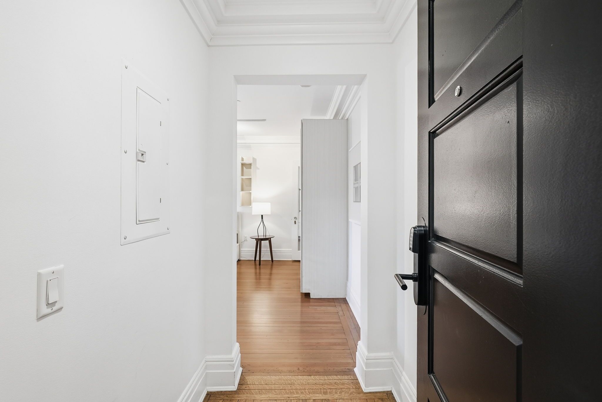 A hallway with a wooden floor, white walls, a black door, and a small table with a lamp.