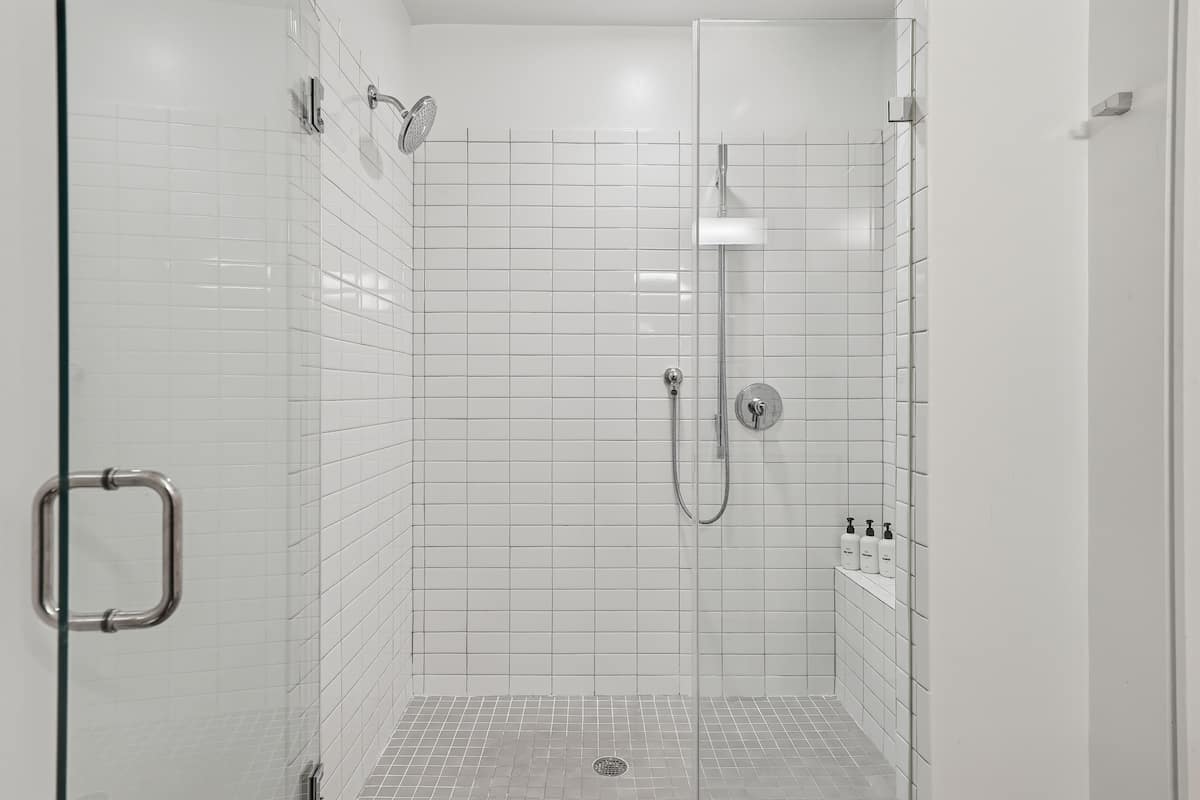 A glass-enclosed shower with white tiles, a handheld showerhead, a wall-mounted faucet, and a shelf with toiletries.