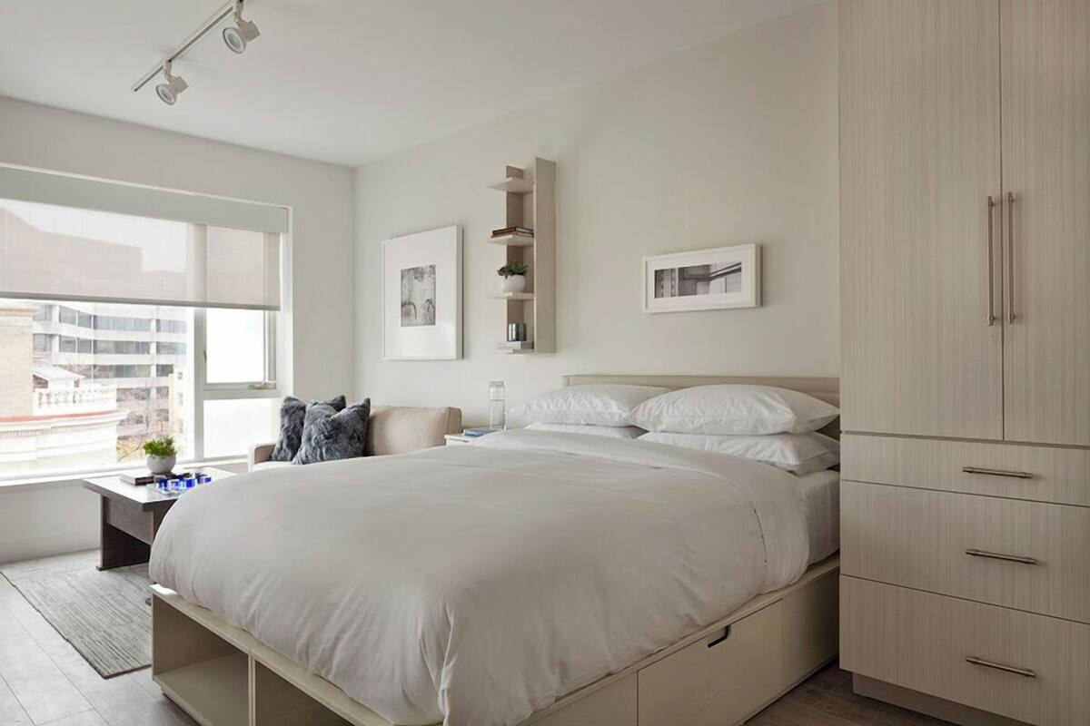 A modern bedroom with a large bed, a nightstand, a wardrobe, and a window with a city view.