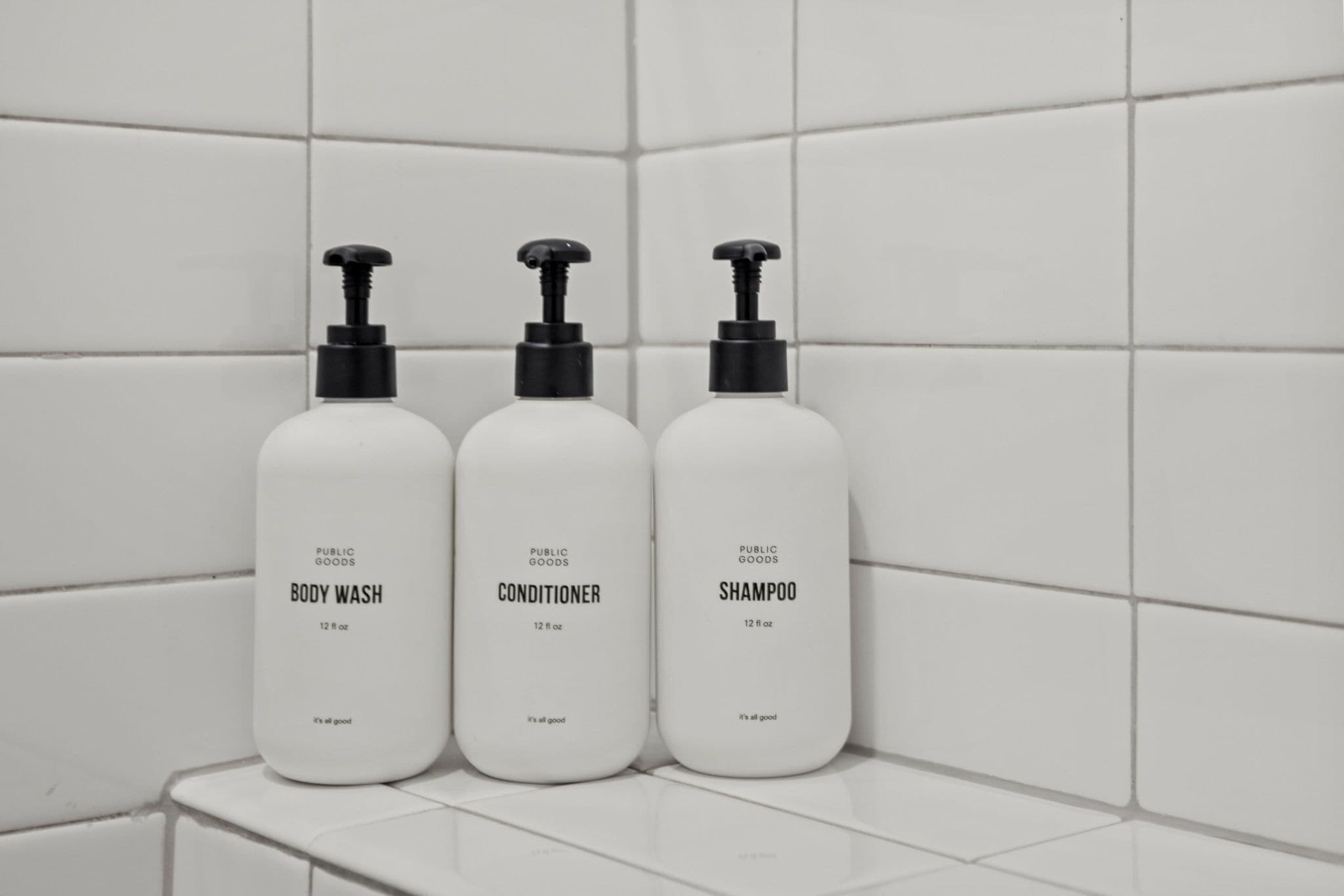 Three white bottles of body wash, conditioner, and shampoo on a white tiled surface.
