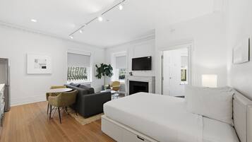A modern bedroom with a white bed, a grey sofa, a small dining table, and a TV mounted above a fireplace.