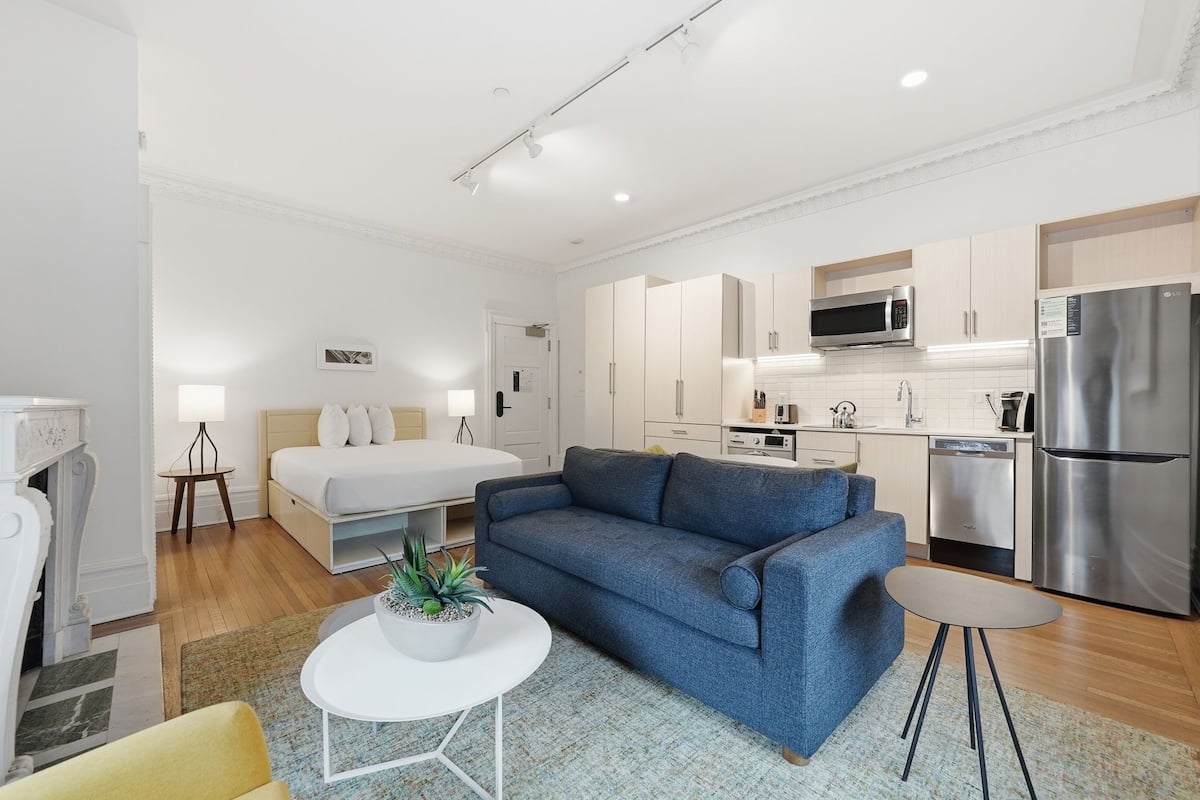 A modern apartment with a bed, sofa, small table, and kitchenette.