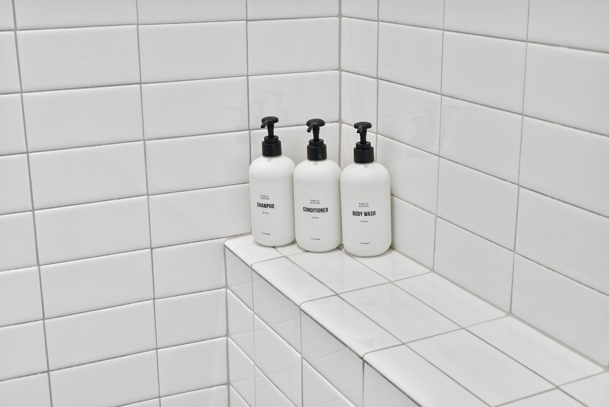 Three white bottles of shampoo, conditioner, and body wash on a white tiled shelf.