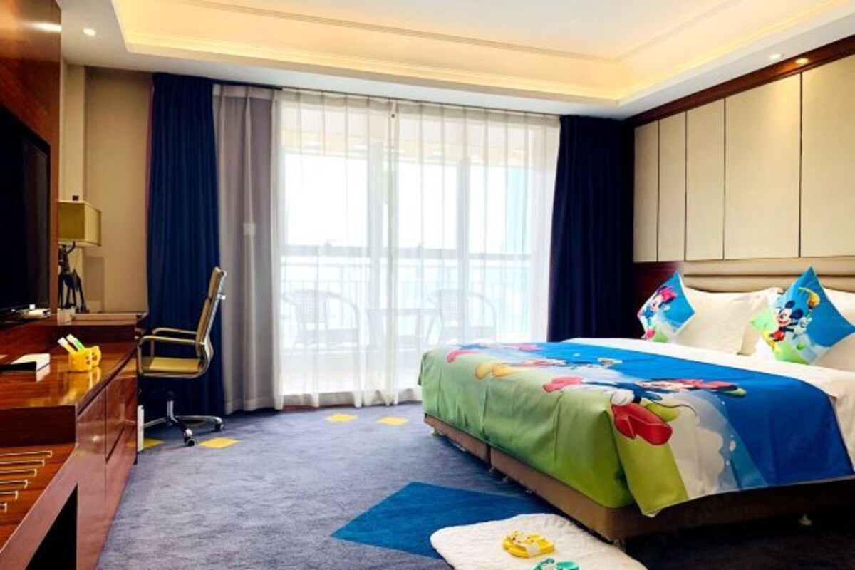 Una camera d'albergo con un letto, una scrivania, una sedia e ampie finestre con tende.