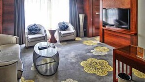 Een hotelkamer met een grote flatscreen televisie, twee fauteuils met blauwe kussens, een ronde salontafel en een kamerbreed tapijt met dessin.