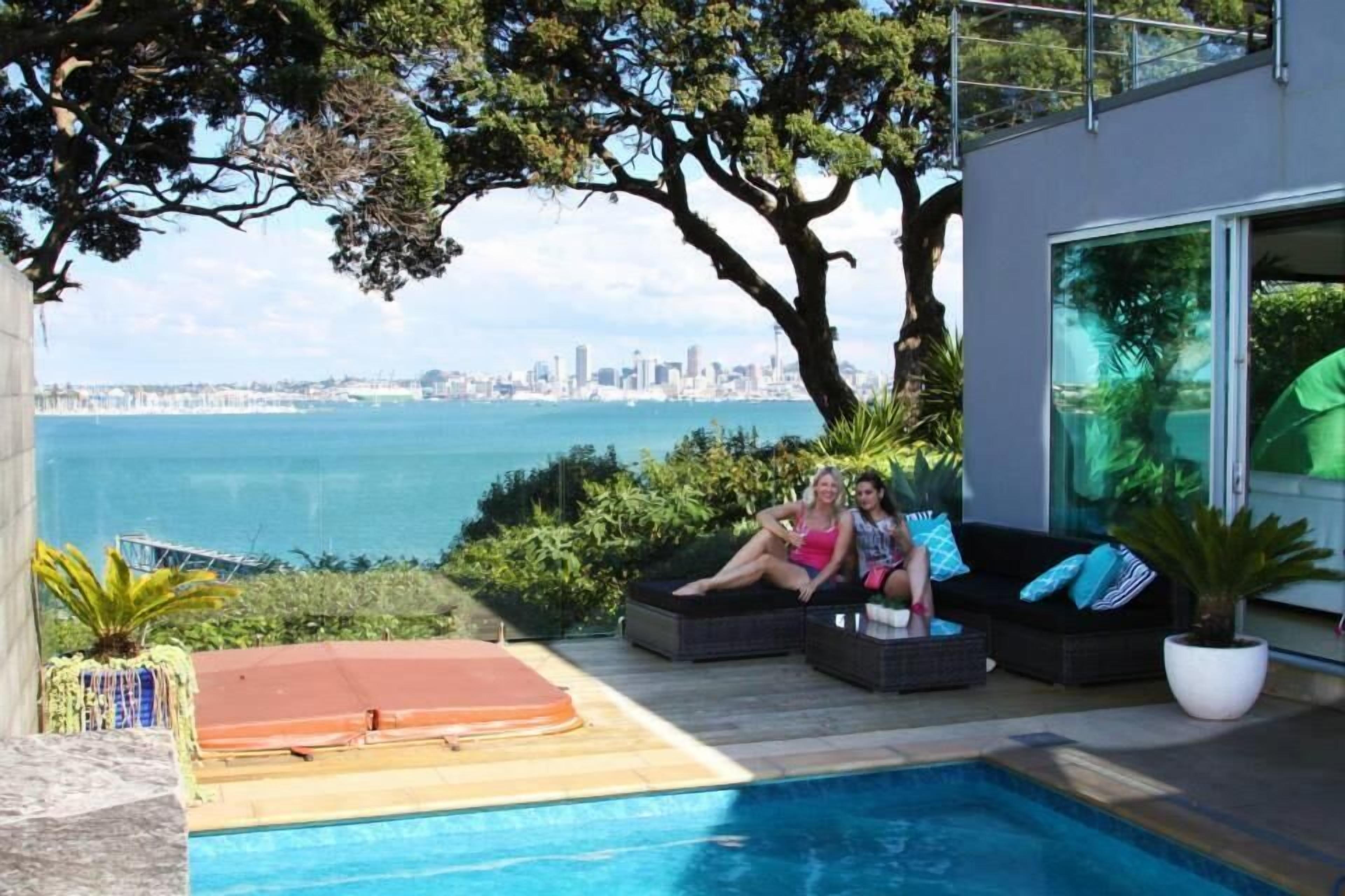 A poolside area with two people relaxing on a sofa, a view of the city skyline, and a large window overlooking the pool.