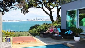 A poolside area with two people relaxing on a sofa, a view of the city skyline, and a large window overlooking the pool.