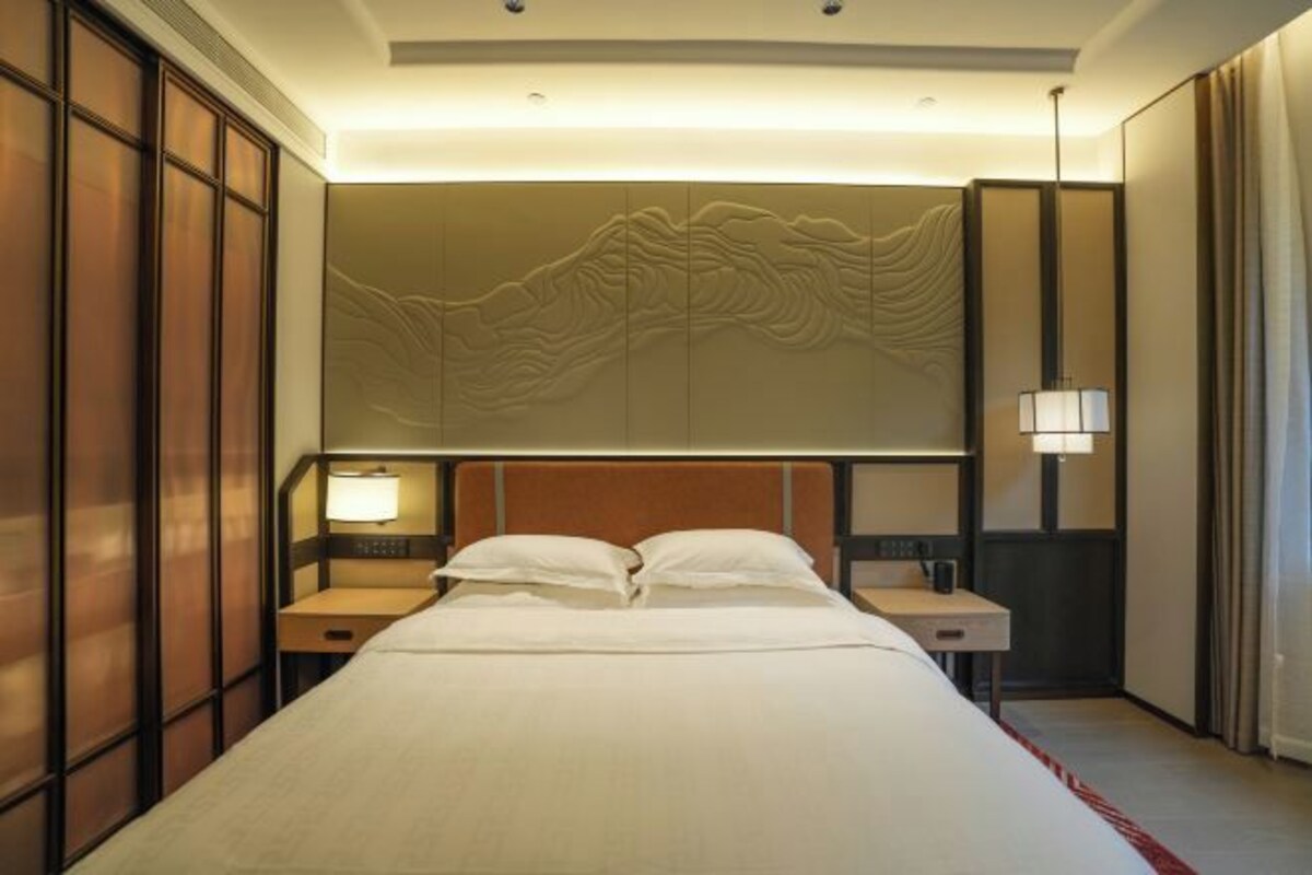 Una habitación de hotel con una cama grande, paneles de madera en las paredes, cabecera con diseño de olas, dos mesitas de noche con lámparas y una alfombra a rayas rojas.