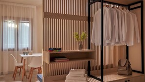 A modern hotel room with a wooden slatted wall, a dining area with a table and chairs, a shelf with books and a vase, and a hanging wardrobe with clothes and a handbag. - "Votsalo" deluxe studio - Studio apt / Sea view /Shared Pool / Sleeps 3 (Malevizi)