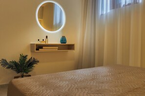 A bedroom with a bed, a bedside plant, a wall-mounted shelf with books and a vase, and a round mirror with lighting.