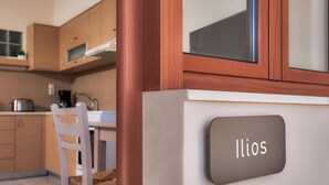 A modern kitchen with wooden cabinets, a white chair, and a sign reading 'Ilios.' - "Ilios" apartment - 1 Bedroom Apt / Sea view /Shared Pool / Sleeps 4 (Malevizi)