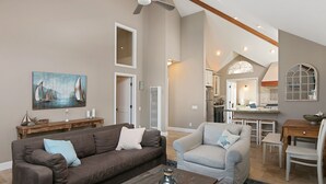 A spacious living room with a brown sofa, a grey armchair, and a wooden coffee table. There is a painting of sailboats on the wall, and a kitchen area is visible in the background.