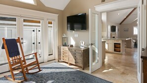A spacious room with a large window, a wooden rocking chair, a dresser, and a flat-screen TV mounted on the wall.