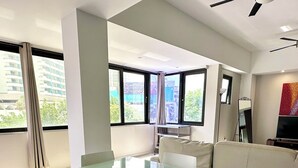 A modern living room with a glass dining table, white chairs, a beige sofa, and large windows offering a city view. - Modern 2BR Apartment Steps from La Concha & the Beach – Prime Condado (Santurce)