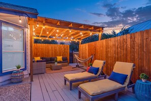 Un patio al aire libre con terraza de madera, una zona de estar techada con luces de cuerda, una hamaca y dos sillones de descanso.
