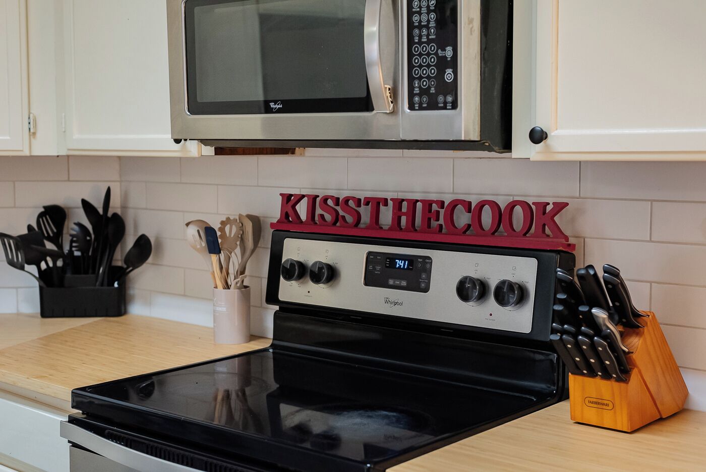 A kitchen with a Whirlpool stove, a microwave above it, and a sign that reads "KISS THE COOK".