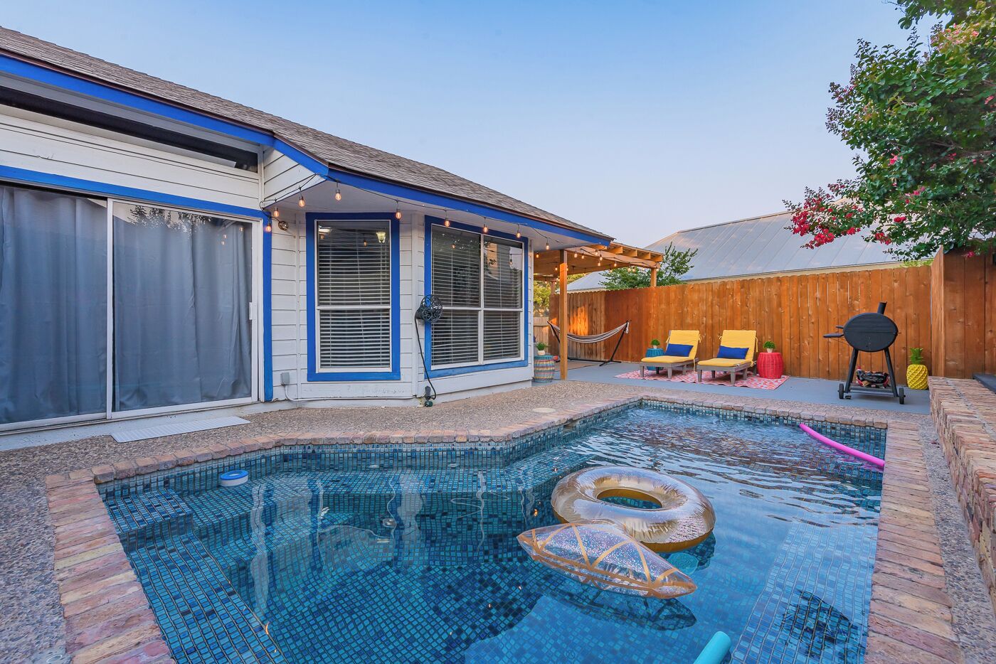 A backyard with a swimming pool, a covered patio area, and a wooden fence.