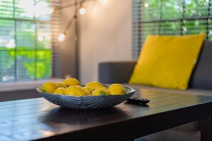 Un cuenco de limones sobre una mesa de madera, con un control remoto a un lado, en una habitación con una ventana y un cojín amarillo.