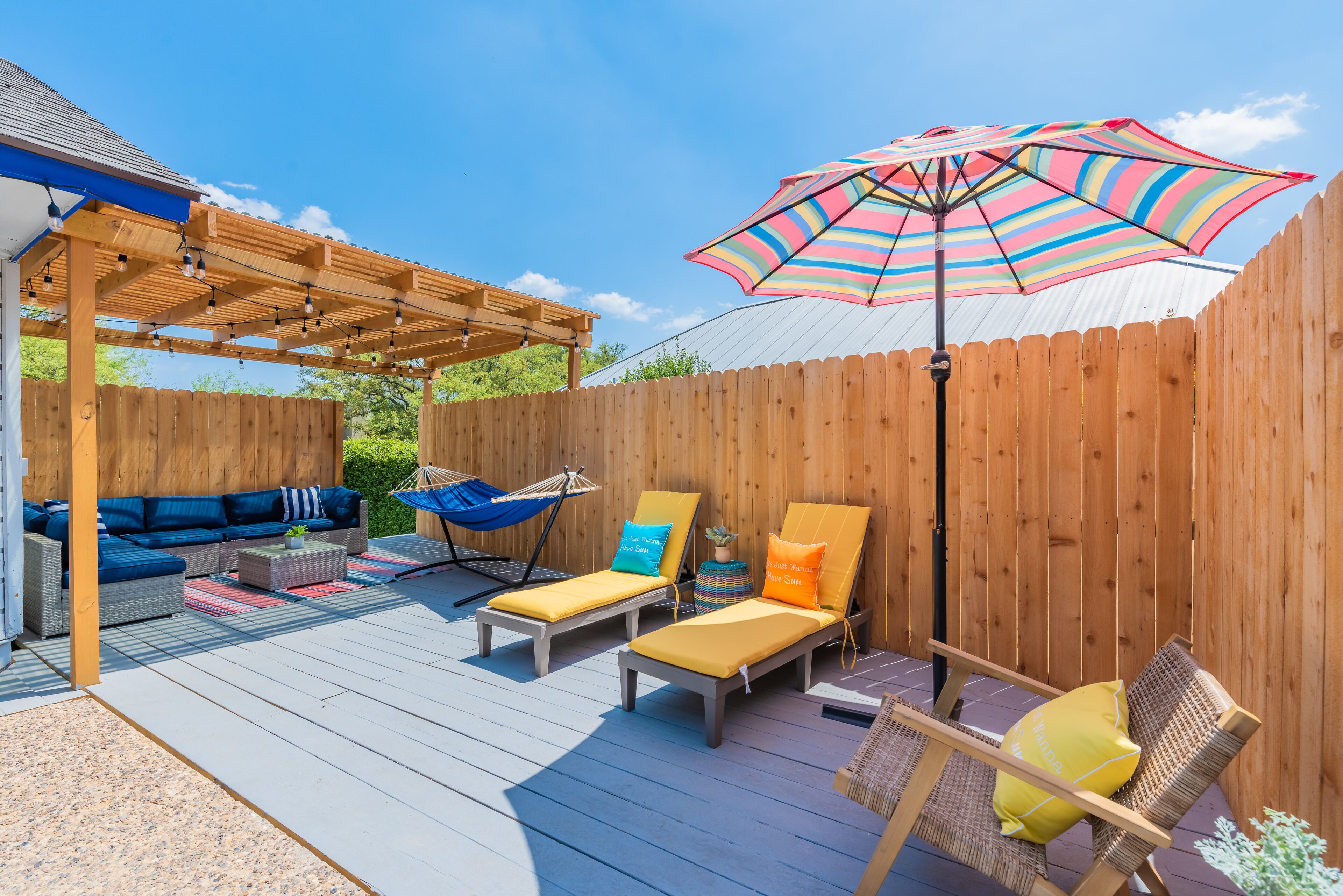 A wooden deck with a covered seating area, including a sofa and two lounge chairs, under a colorful striped umbrella.