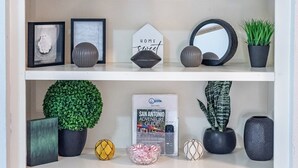 Una estantería con objetos decorativos como letras, ilustraciones enmarcadas, un letrero que dice 'Hogar Dulce Hogar', una guía de aventuras por San Antonio y varias plantas en macetas.