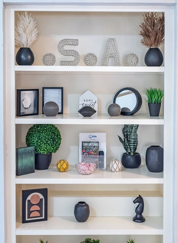 Una estantería con objetos decorativos como letras, ilustraciones enmarcadas, un letrero que dice 'Hogar Dulce Hogar', una guía de aventuras por San Antonio y varias plantas en macetas.