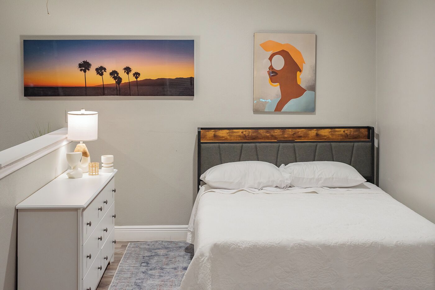 Een slaapkamer met een bed, een nachtkastje, een lamp, een schilderij van een zonsondergang en nog een schilderij van een persoon.