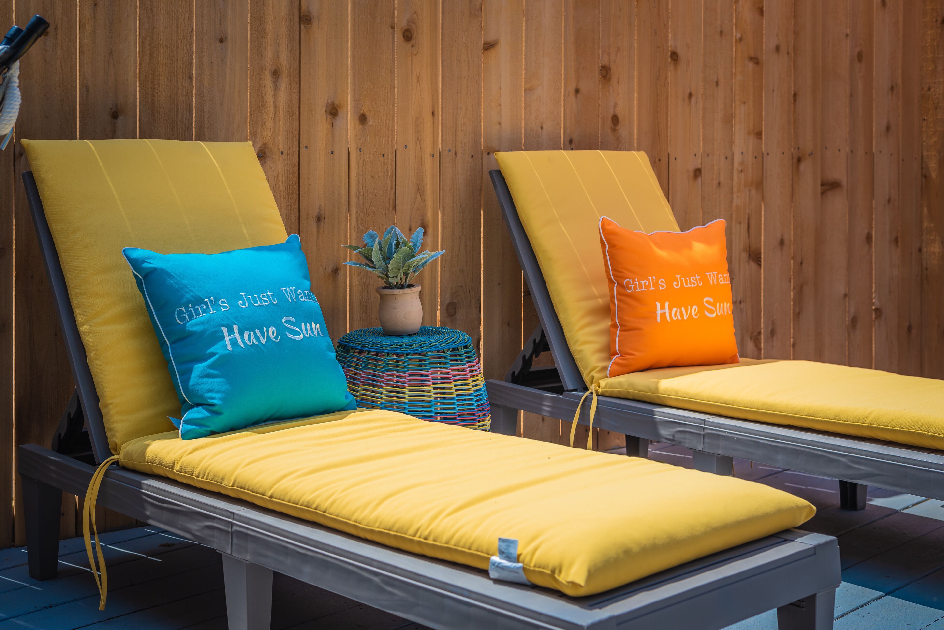 Deux chaises longues avec des coussins et des oreillers jaunes ornés de motifs, une petite plante en pot et un mur en bois en arrière-plan.