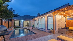 Patio trasero con piscina, parrilla y área de terraza cubierta con luces colgantes.