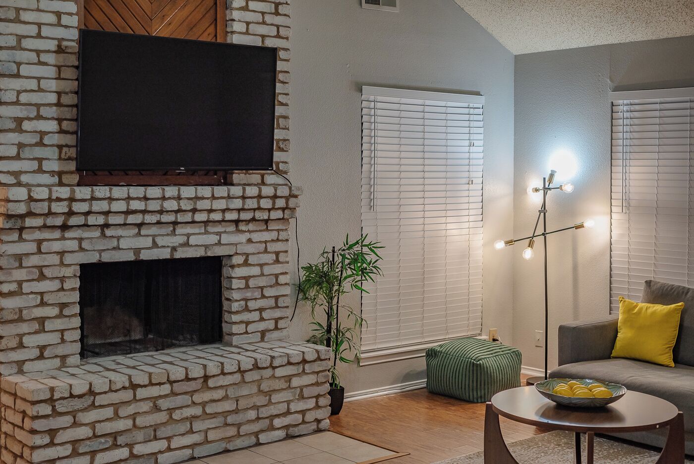 Una sala de estar con chimenea de ladrillo, un televisor de pantalla plana montado encima y una lámpara de pie moderna.