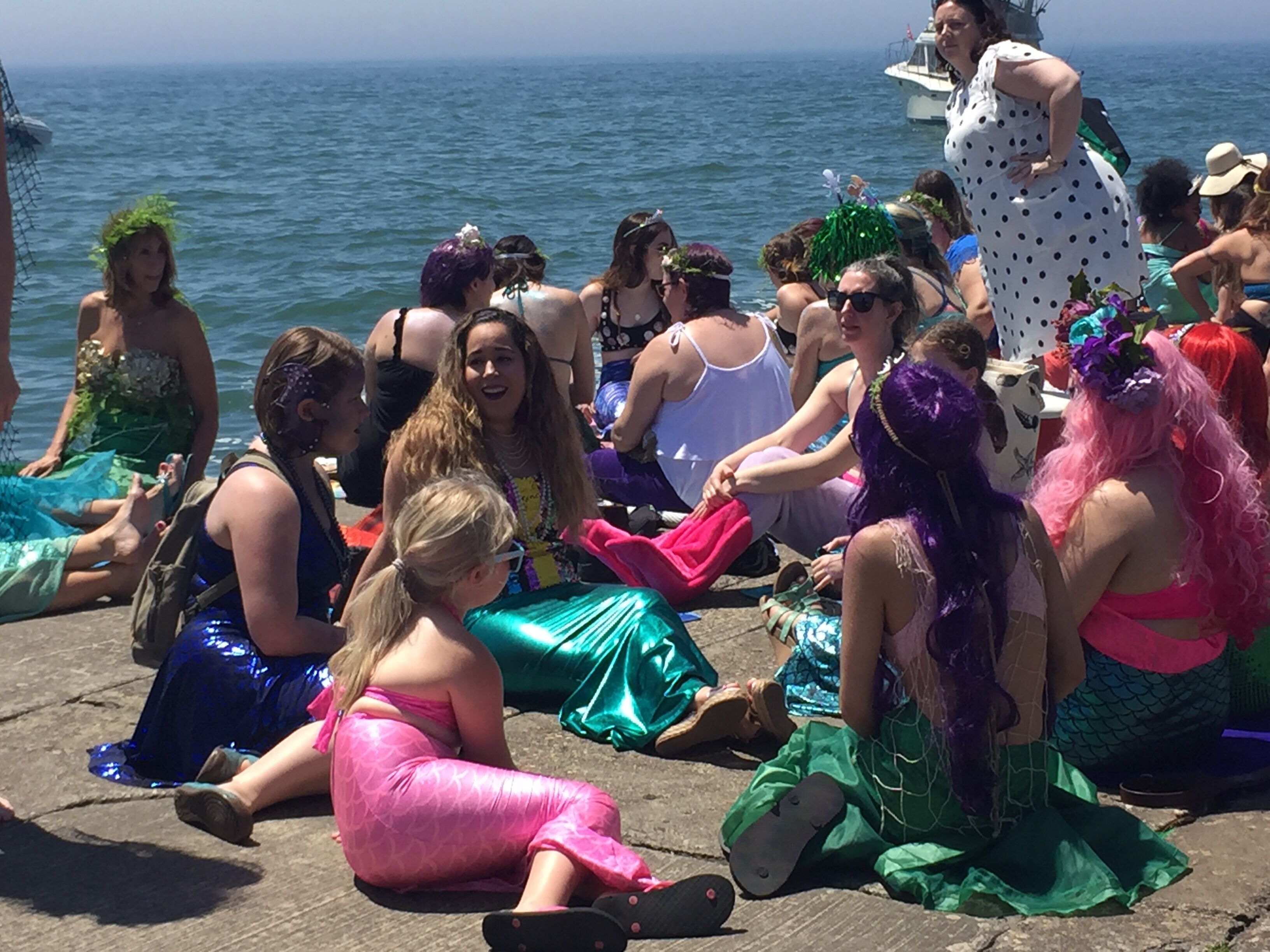 Een groep mensen zit op een houten steiger aan de zee, gekleed in kleurrijke kostuums.