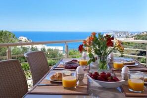 Une table de petit-déjeuner avec des croissants, des fraises, du jus d’orange et un vase de fleurs, offrant une vue panoramique sur la côte.
