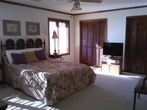 Un dormitorio con cama, televisor sobre un mueble, ventana con persianas y ventilador de techo.