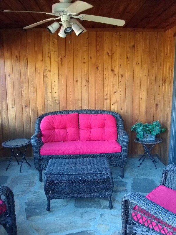A covered patio area with a wicker sofa and ottoman, a small table, and a ceiling fan.