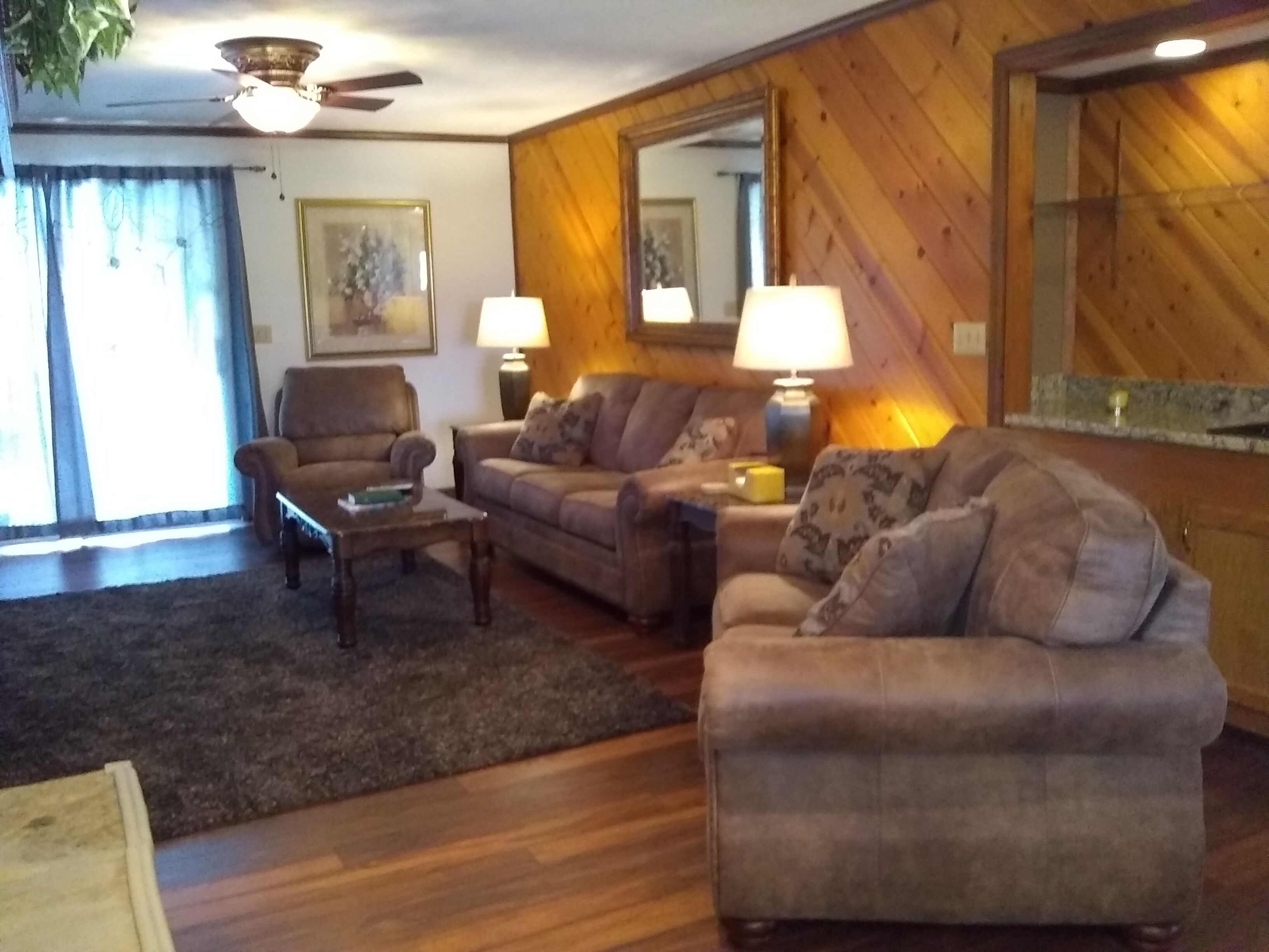 A cozy living room with a sofa, armchair, and coffee table. There is a ceiling fan, a lamp, and a framed picture on the wall.