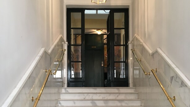 Un escalier en marbre, agrémenté de rampes dorées, mène à une porte sombre ornée de panneaux de verre.