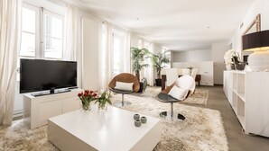 Un salon moderne avec un téléviseur à écran plat, une table basse blanche, deux fauteuils et un canapé. On y trouve également des plantes en pot et une grande fenêtre avec des rideaux.