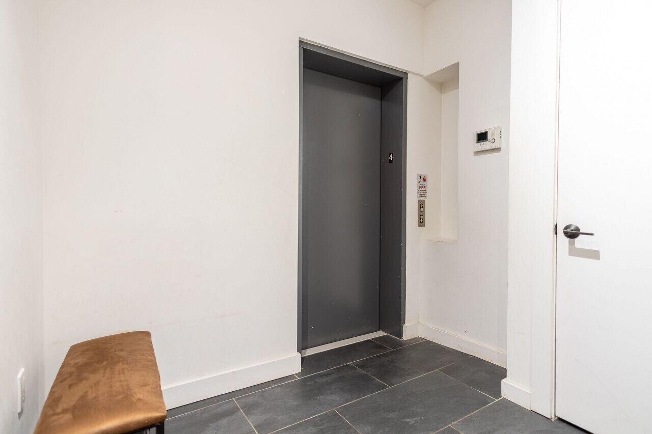 A hallway with a grey elevator, a bench, and a white door with a fire alarm.
