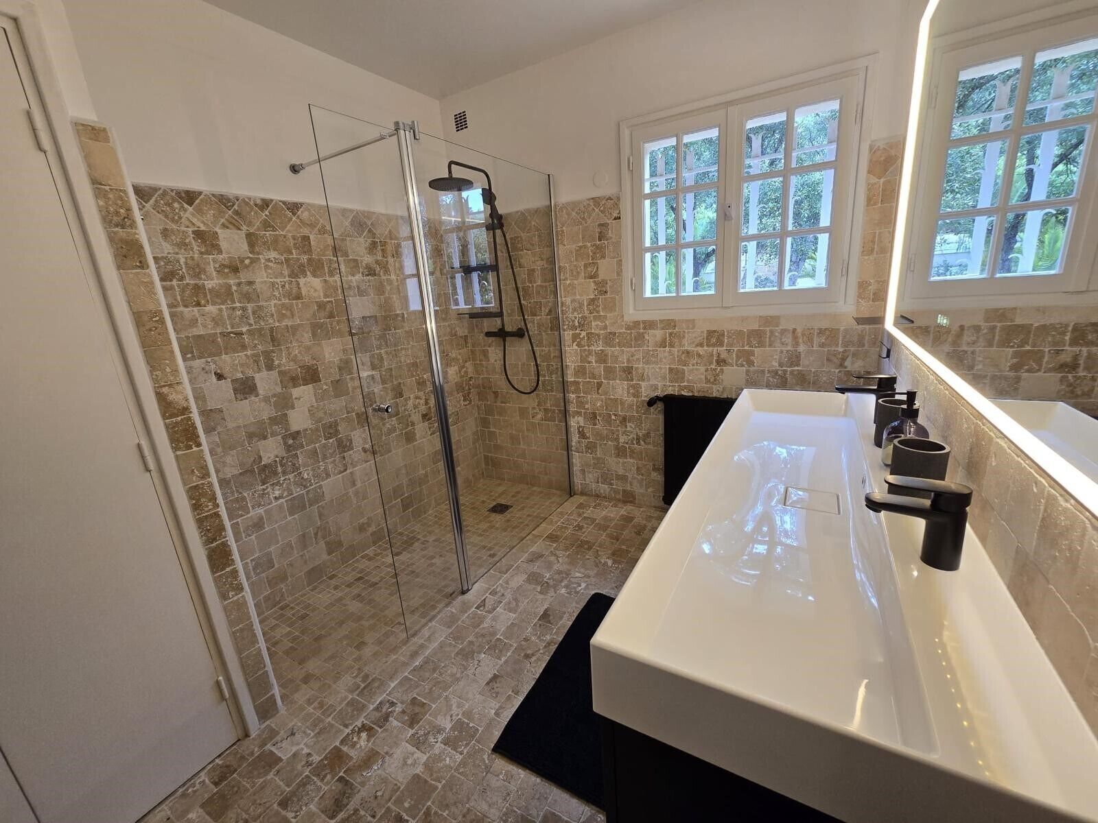 Et moderne badeværelse med et glasafskærmet brusebad, en stor hvid vask og et vindue med naturligt lys.