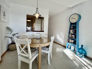 Un coin repas comprenant une table ronde en verre, des chaises blanches, un luminaire suspendu, une bibliothèque remplie de livres et une horloge décorative.